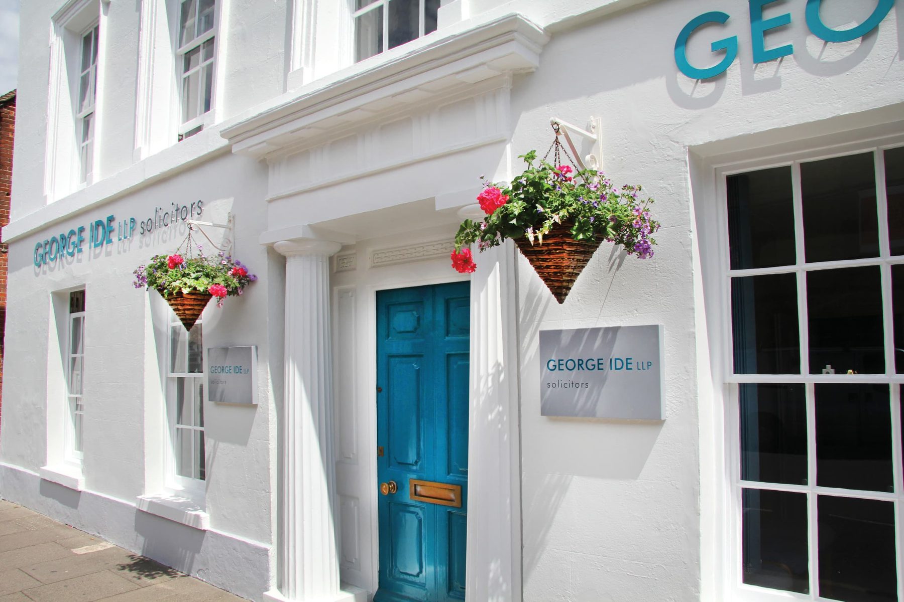 The George Ide LLP shopfront at 52 North Street, Chichester: a white-rendered Georgian-style facade with a striking teal-painted front door, twin slate-grey signage plaques reading GEORGE IDE LLP solicitors, two hanging baskets of pink and purple geraniums above the door, and large teal cut-out GEORGE IDE lettering mounted high on the wall.